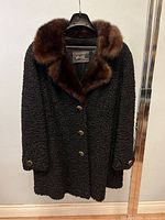 Full front view of the vintage Persian lamb fur coat showing the rich black curled lamb wool and brown mink collar, button front closure, and long sleeves.