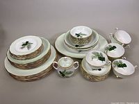 Full set displayed on table showing stacked plates, sugar bowl, teacups, and open vegetable bowls with green botanical pattern and gold rims.
