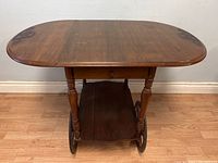 Front view of the tea trolley showing the drop leaf top, turned legs, drawer, two large wheels, and lower shelf.