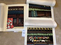 Three woodblock prints stacked with protective acid-free paper, showing cut-edge of prints with colorful storefront and fan scenes.