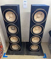 Front view of both tower speakers, each with four exposed 7-inch drivers, black wooden enclosures, 13-inch square bases