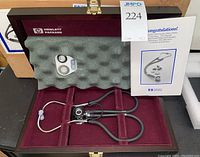 Wooden presentation case open with stethoscope in foam-lined interior, spare parts tray and manual visible
