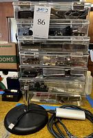 Stack of seven clear plastic bins with assorted microphone and audio equipment parts visible inside, and a small desktop microphone stand with USB cable and silver microphone component in front.