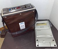 Brown carrying case fitted with portable Sony Betamax videocassette recorder next to silver carrying sleeve, and stack of blank videocassettes