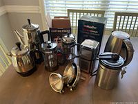 View of assortment on table showing Farberware coffee pots, French press coffee presses, coffee canister, electric kettle, coffee funnel, manual grinder, and boxed Mueller SuperGrind coffee grinder.