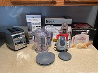 Photo of all kitchen appliances on countertop including Ninja Master Prep with parts, toaster, Donvier ice cream maker, and Once For All mandolin slicer with boxes visible behind.
