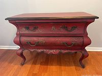 Front view of the red Bombay style chest showing two drawers with Asian design and ornate metal handles, standing on cabriole legs.