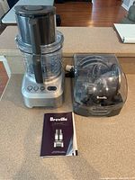 View of Breville The Sous Chef food processor with work bowl, black lid with feeding tube, accessory container with discs, and user booklet on a kitchen counter.