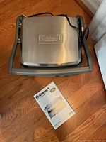 Cuisinart stainless steel griddler closed on hardwood floor with instruction manual in front.