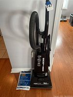 Full view of the Eureka Victory vacuum with black hose and handle, standing upright with the package of vacuum bags beside it on the floor.