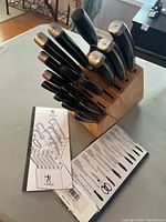 View of the cutlery set upright in the wooden block on a table, showing multiple knives with black handles and the JA Henckels logos on the ends.