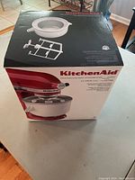 Photo of KitchenAid Ice Cream Maker attachment box showing the unit attached to a red KitchenAid stand mixer.