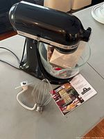 Black KitchenAid Artisan stand mixer with glass mixing bowl inside and wire whip attachment placed beside it on the counter, along with instruction manuals.