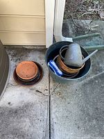 Photo showing stacked terracotta and glazed pots with a blue scrubbing brush inside a large glazed dark pot, placed on concrete surface next to house exterior.