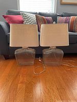 Pair of table lamps placed side-by-side on hardwood floor in front of sofa with cushions, showing full view of lamps with clear glass bases and beige fabric shades.
