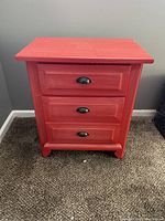 Front view of red wood nightstand with visible three drawers and black metal handles, showing overall condition and style.