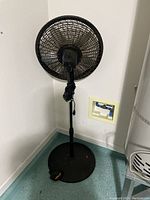 Photo of a black tall floor fan with protective grille and three blades, positioned in a corner near a water heater and laundry area.
