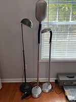 Three floor lamps standing side by side near a window, showing black lamp and two silver lamps with adjustable arms.