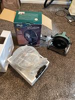 Hunter Century ventilator fan box with fan parts in styrofoam and plastic wrapping alongside small portable green metal fan on carpet.