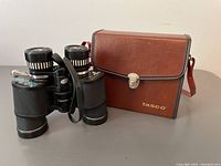 Full view of vintage Tasco binoculars next to their brown leather carrying case with shoulder strap and metal clasp.