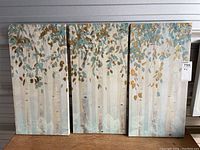 Three matching vertical canvas prints depicting birch trees with blue and gold leaves, arranged side by side on a shelf.