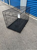 Black folding metal dog crate with single front door and plastic tray inside shown fully assembled outdoors on pavement.