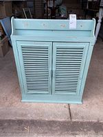 Front view of the light blue painted wooden medicine cabinet with shutter style double doors and some wear marks on the top shelf area.