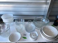 View of assorted glass bowls, pie plates, ceramic mixing bowls, Pyrex measuring cups, and baking dishes with woven holders displayed on a white surface.
