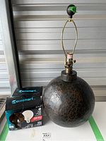 Round leather covered table lamp standing on a white surface with two boxed Ecosmart LED bulbs placed next to it. The lamp features a cracked textured leather surface on a spherical base and a brass lamp holder with a curved support for the lampshade.