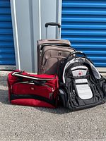 Brown Delsey carry-on suitcase, red Swiss Gear tote bag, and black Swiss Gear backpack grouped together on pavement outdoors against blue storage unit doors.