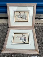 Pair of framed prints showing stylized mounted figures on horses, arranged vertically outdoors on concrete ground near blue metal background