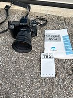 Minolta Maxxum HT Si camera with zoom lens attached, instruction manual, and an arm strap laid out on the ground