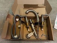 Top view of Moen Brantford widespread faucet components laid in box. Includes spout, hot and cold handles, hoses, and mounting screws.