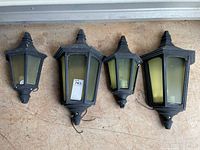 Four outdoor coach lights arranged on ground showing two large and two small units with discolored yellowed glass panels and wrought iron frames.