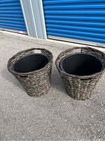 Two brown resin woven-style planters with handles, placed outdoors on pavement near blue storage doors. Each planter has a removable black plastic inner pot.