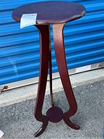 Full view of the three-legged wooden plant stand with round top and curved legs placed outside on pavement.