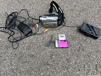 Overview of all three items: Panasonic camcorder with charger and cables, pink Olympus camera, and black Sanyo Walkman.
