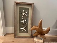 Photo showing 3 barnboard framed starfish on the left and a standalone carved wooden starfish sculpture on the right against a light-colored wall on floor.