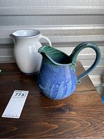 Photo showing both ceramic pitchers side by side, white one with chip and signed Jane and blue-green glazed one.