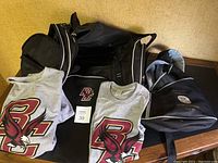 Photo showing Boston College grey sweatshirt, T-shirt with eagle logo, large black gym bag, and navy blue 1959 Reunion backpack on a wooden surface.