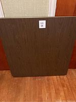 Full view of square collapsible bridge table top with wood grain finish leaning vertically against a wall.