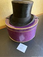 Black tall top hat with a slightly shiny textured surface and a black ribbon band around the base of the crown, resting on a purple hat box with gold trim. Both items show signs of wear.