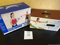 Photo showing two boxed exercise products: BFIT Gear Arm Sculpting System and Natural Fitness Inc. Soft Weighted Balls, with lot number card in front.