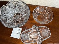 Top view of three cut glass decorative items on a table: large cut glass bowl, small floral etched bowl, and handled decorative basket with damaged handle.