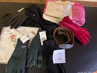 Photo showing multiple pairs of ladies winter gloves in gray, black, dark brown, and bright pink velvet. Also shows a pink and cream winter scarf, vintage brown leather belt with round buckle, and a set of unopened embroidered handkerchiefs.