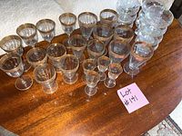 Full lot of various sized glasses arranged on a wooden table showing stack of clear champagne glasses on right and multiple rows of gold rimmed glasses.