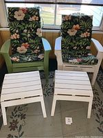 Green and white plastic Adirondack chairs with floral cushions and two white small tables or footrests in front