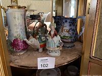Porcelain items grouped on a round table showing the teapot, lady figurine with flowers, Japanese elephant, and large decorative pitcher.