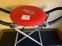 Close-up image showing the red padded seat of the Red Fitness CX machine with logo and folding frame beneath.