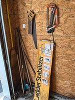 View of ladder stabilizer box and assorted yard tools including broom, saws, and long-handled tools.